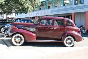 An old car parked in front of the Masonic Building.  Festival goers having lunch in the background.